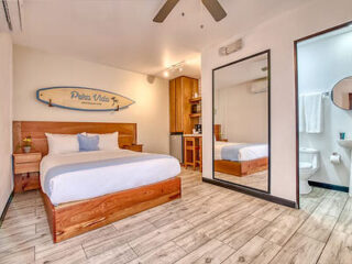 A modern hotel room with a queen-sized bed, a surfboard wall decoration, wooden furnishings, a large mirror, and a small bathroom to the right. The decor is beach-themed with light colors.