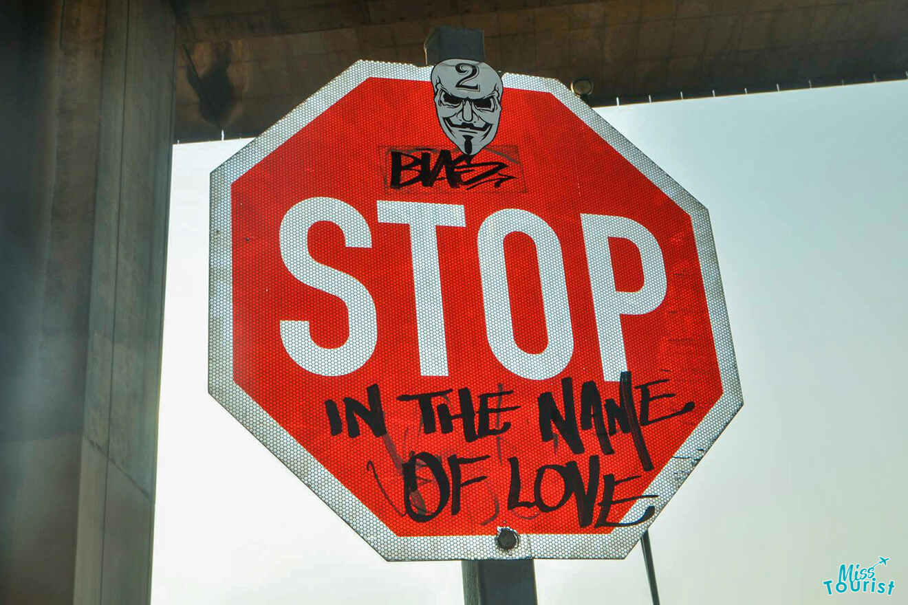 A red stop sign with black graffiti reading "IN THE NAME OF LOVE" below the word "STOP." The sign also features a sticker of a masked face and a tag that reads "Blaz.