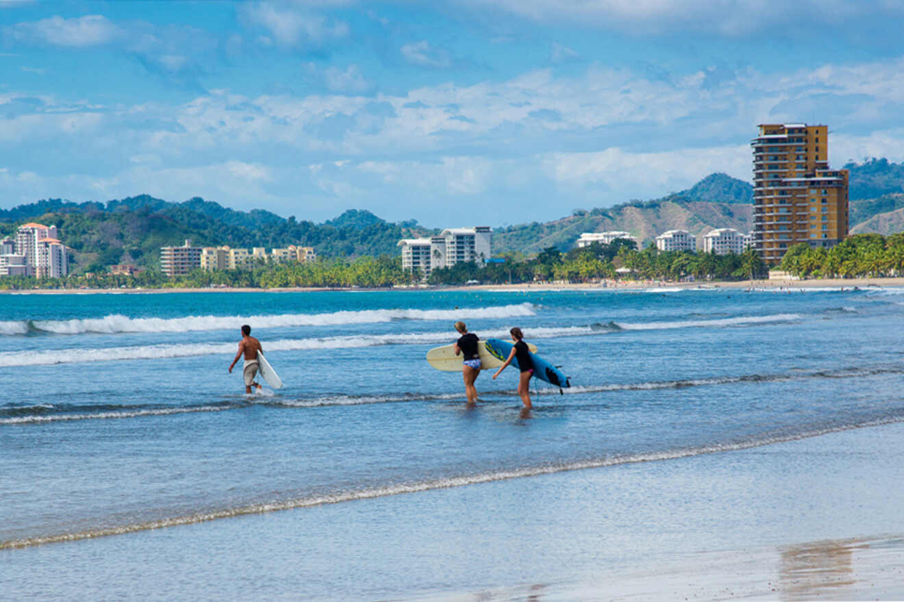 Three people carrying surfboards walk towards the ocean on a beach with buildings and mountains in the background. The sky is partly cloudy.