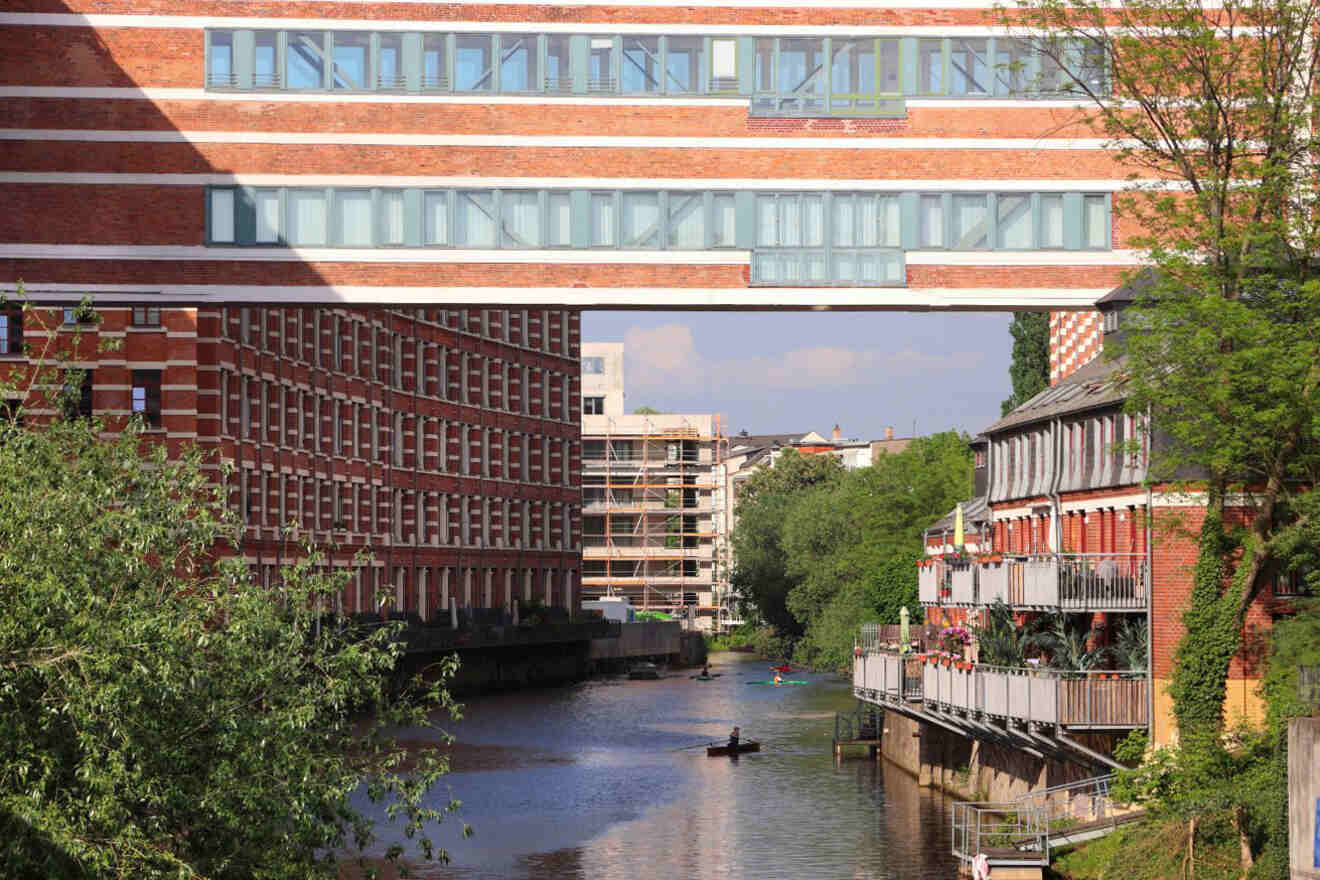Brick buildings and a suspended bridge overlook a canal with a kayaker, tree-lined banks, and residential balconies.