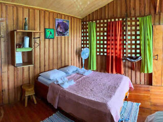 A small wooden cabin bedroom with a double bed, pink bedspread, fan, shelves, and colorful curtains. Walls are wood-paneled with a couple of pictures hanging. A stool and hammock are also visible.