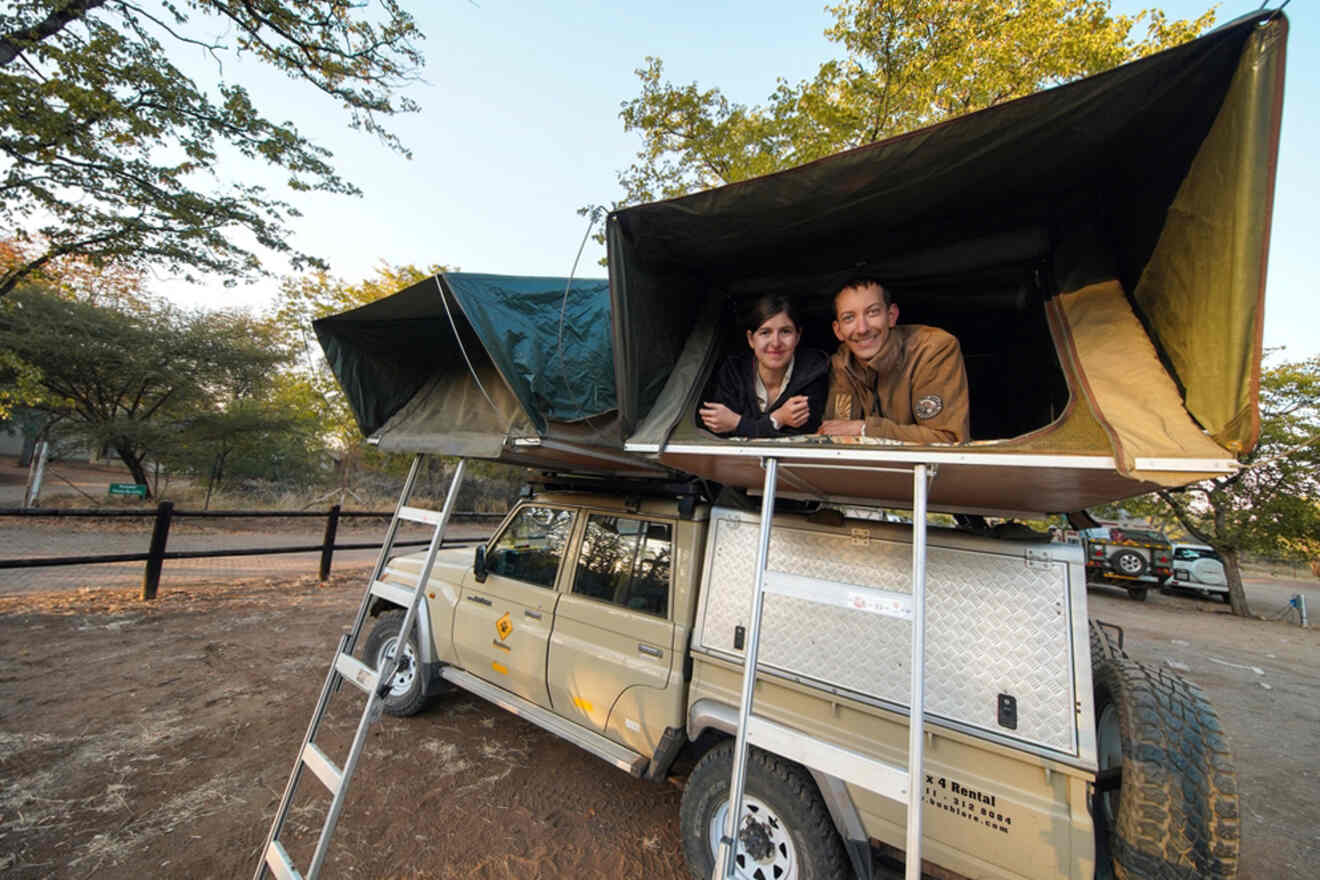 Two people in a rooftop tent atop a parked off-road vehicle with ladders, in a wooded area.
