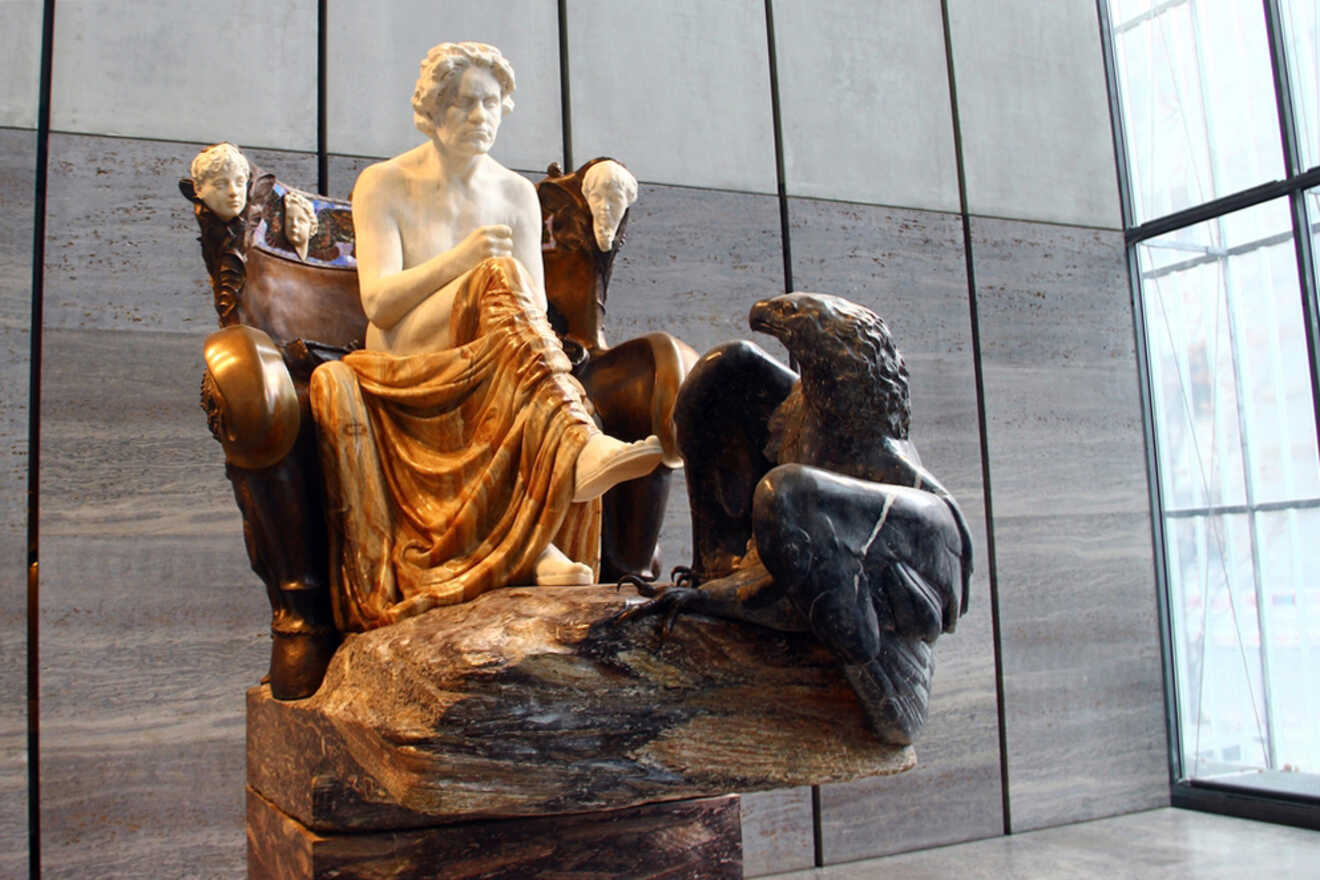 A marble and bronze sculpture depicts a seated figure with his head resting on his hand, alongside an eagle. The background features large windows and a gray wall.