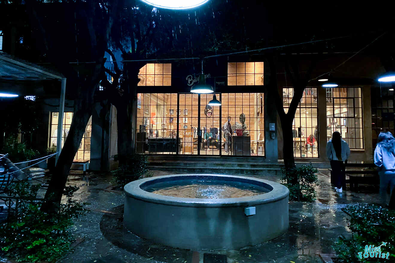 Outdoor area of a building with large glass windows lighting up the interior, showing people inside. A circular water fountain is in the foreground, surrounded by wet ground reflecting night lights.
