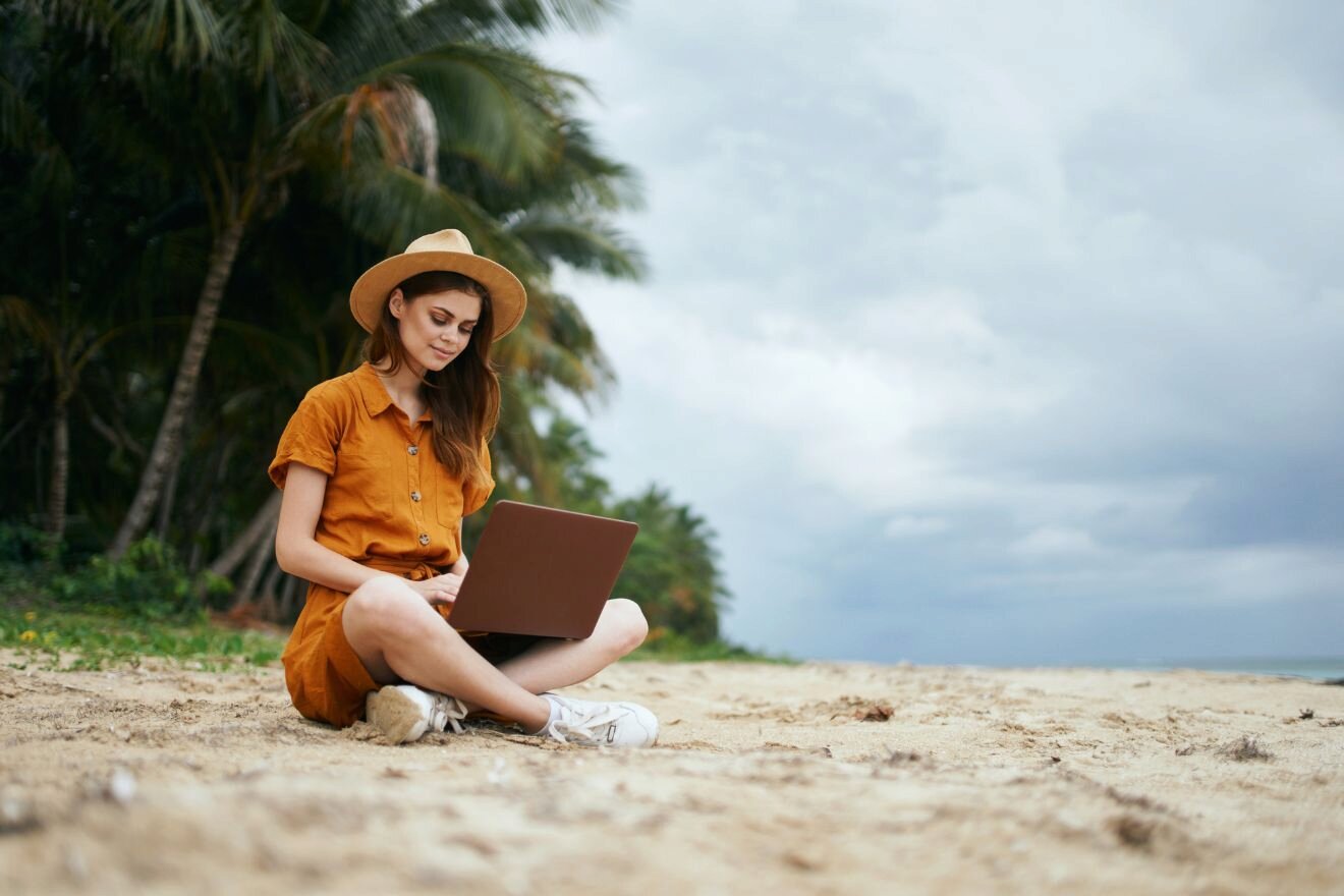 A person wearing a hat and orange outfit sits on the sand near palm trees, using a laptop near the beach under a cloudy sky.