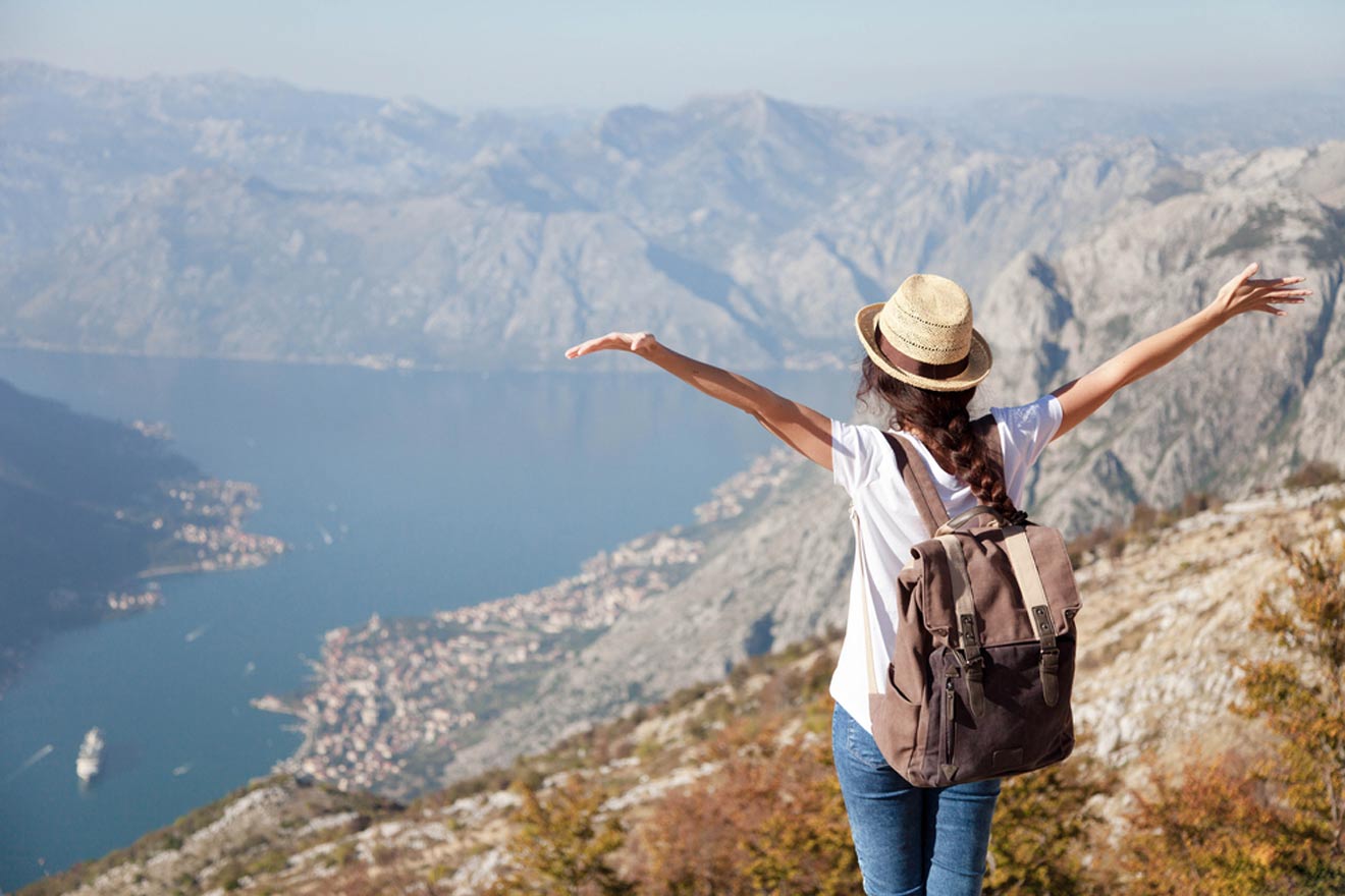 A woman with a backpack and hat stands on a mountain trail, arms outstretched, overlooking a coastal town and body of water below.