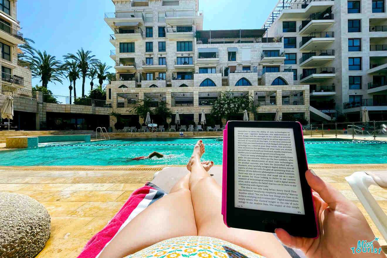 Person reclining on a lounge chair by a pool reading on an e-reader. Buildings and palm trees are in the background.