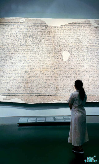 Author of the post in a white dress stands in front of a large ancient manuscript displayed in a glass case at a museum or exhibition.
