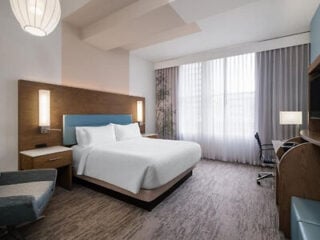 Modern hotel room with a king bed, two nightstands, a chair, desk, lamp, and large window with sheer curtains allowing natural light.