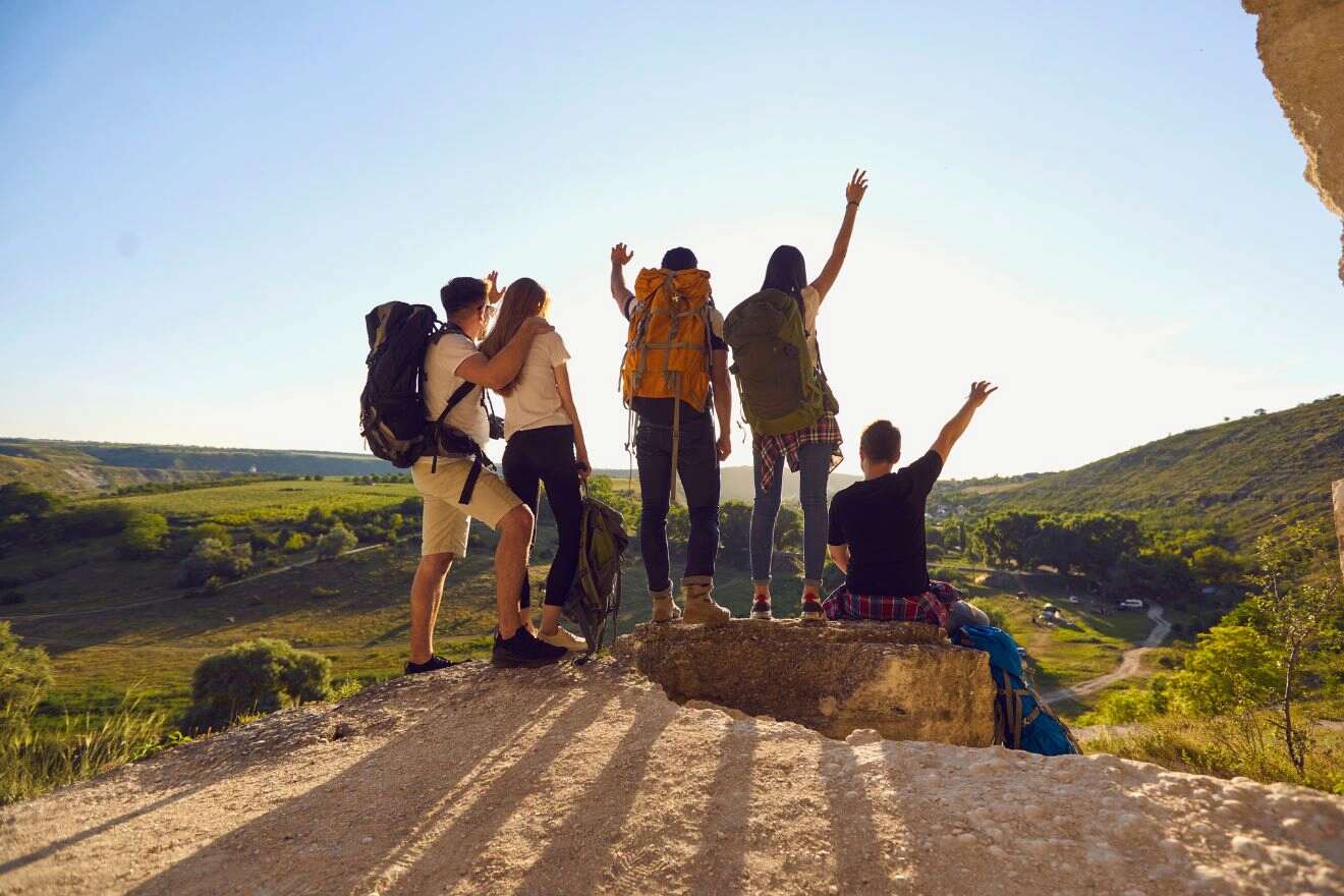 Four hikers standing on a rock ledge, overlooking a scenic valley, with two raising arms in celebration. It is sunny, and they are wearing backpacks.