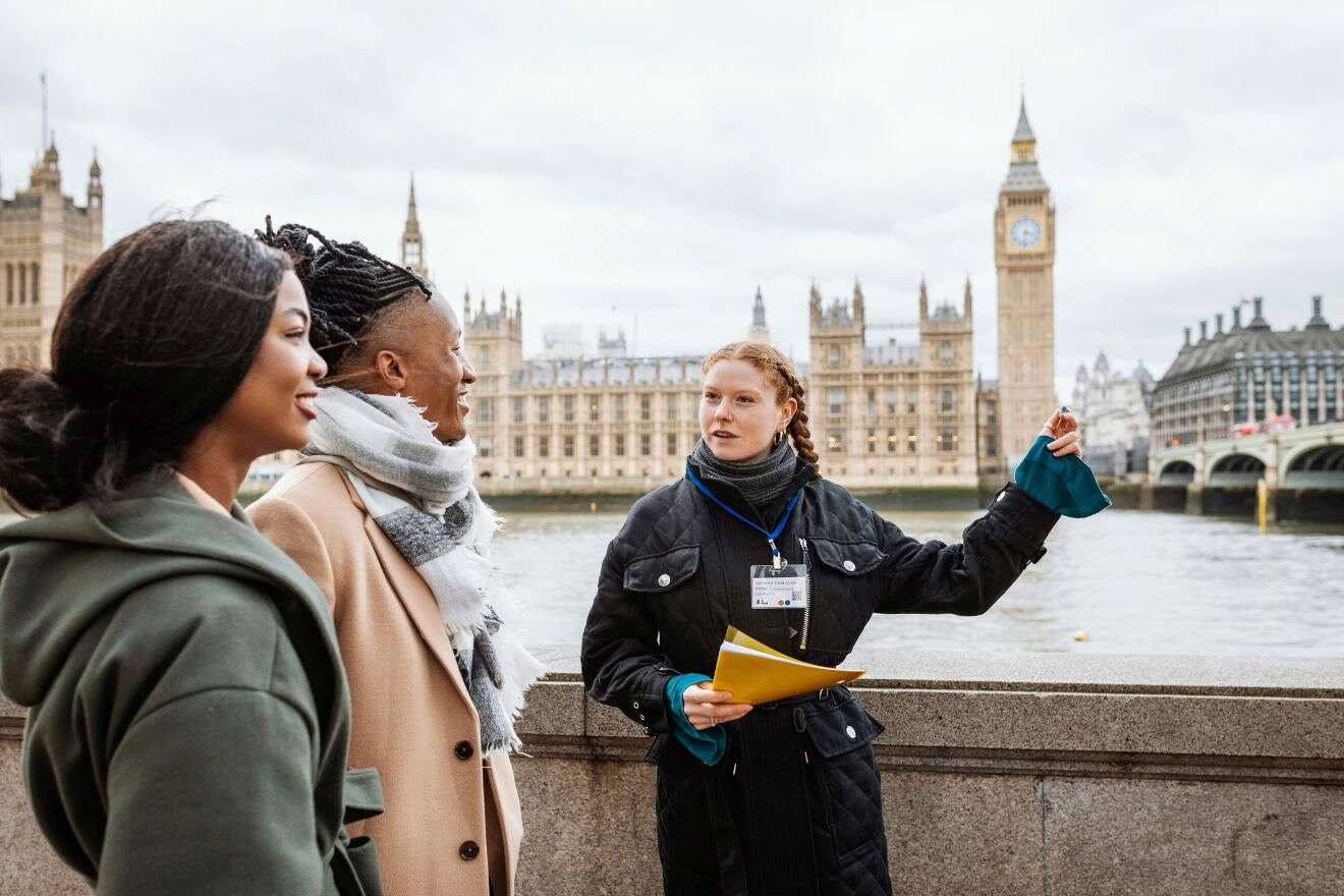 A tour guide gestures while talking to two people in front of the Palace of Westminster and Big Ben in London.