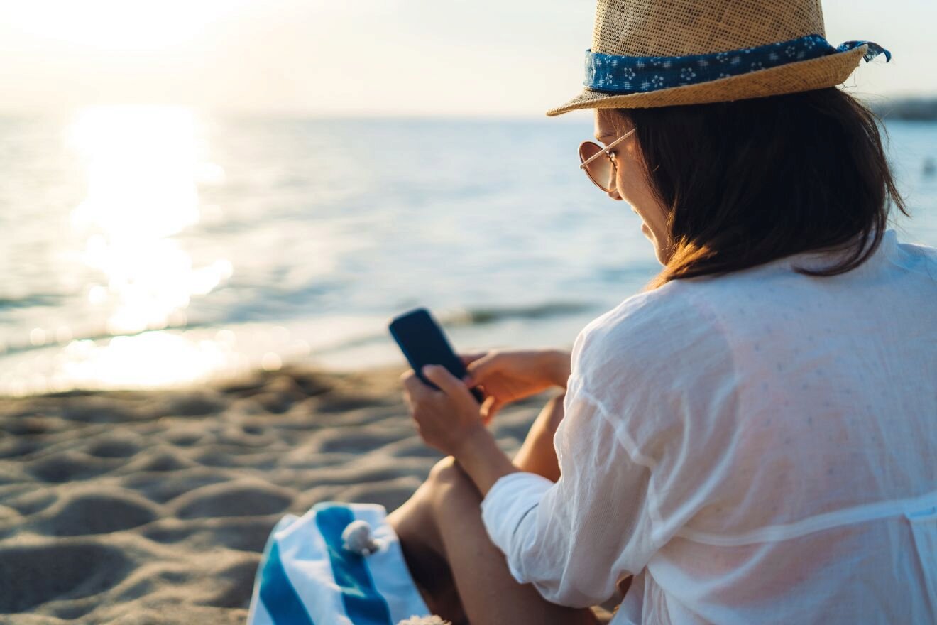 A person wearing a hat and sunglasses sits on a beach, looking at a smartphone. The sun is setting over the ocean in the background.