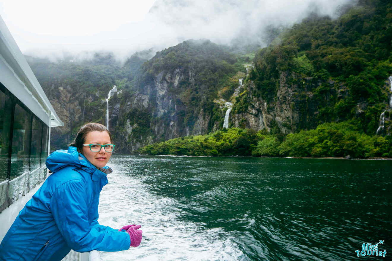 The author of the post in a blue jacket and pink gloves stands on a boat, gazing at misty mountains and waterfalls across a green, forested shore.