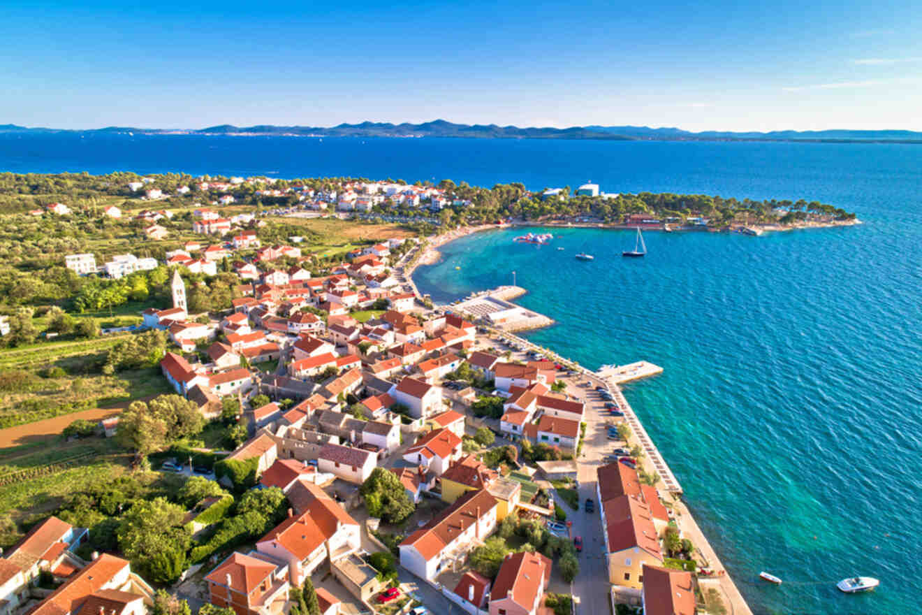 Aerial view of a coastal town with red-roofed houses, a waterfront promenade, and several boats docked in the blue waters of the Adriatic Sea under a clear blue sky.
