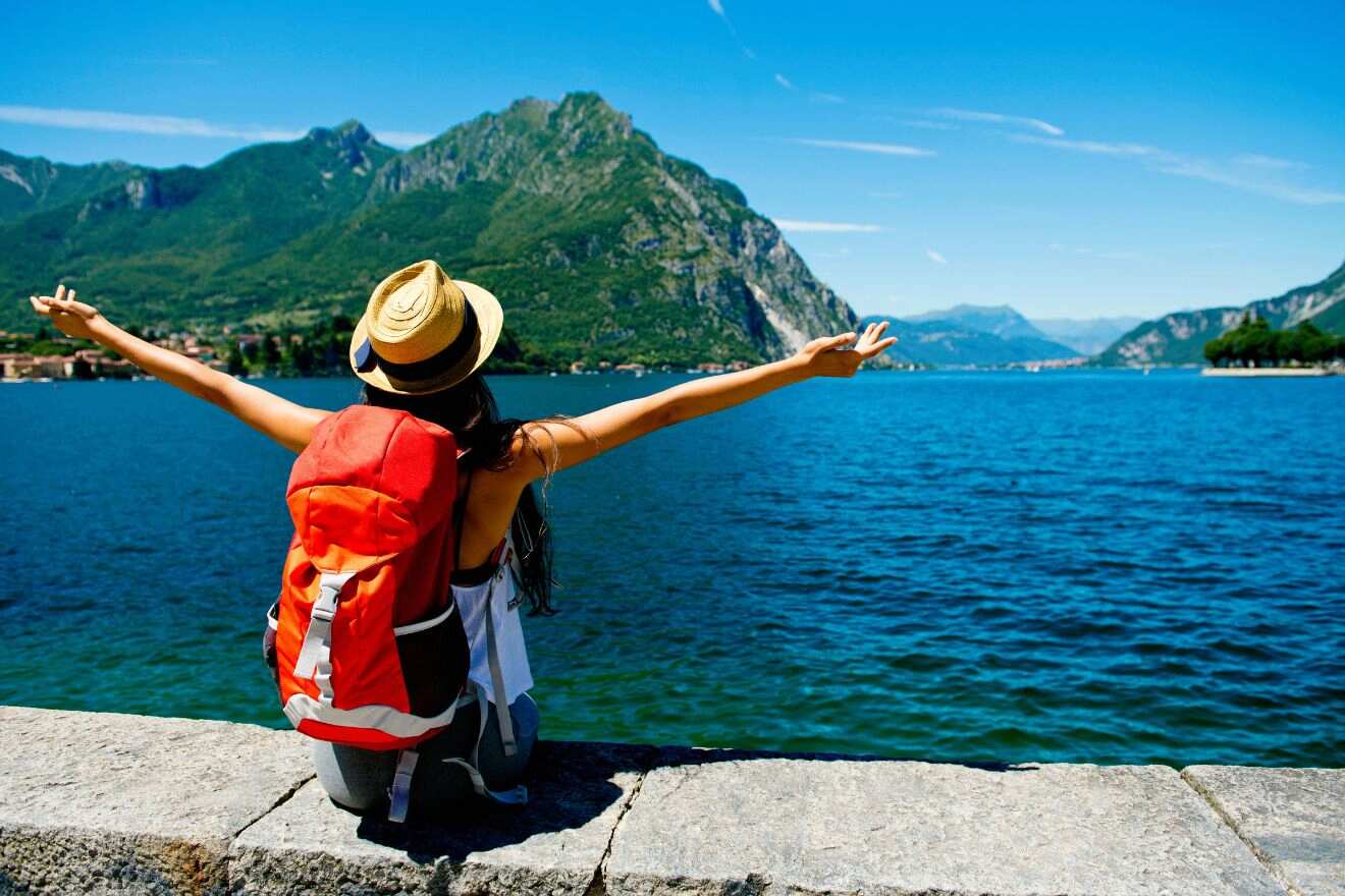 A person with a hat and red backpack sits on a stone wall, facing a large lake surrounded by mountains, with arms outstretched.