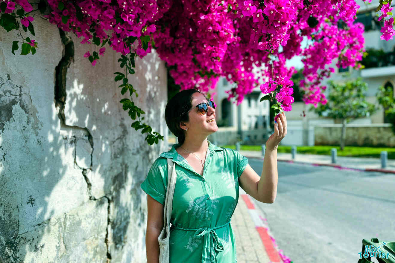 Author of the post in a green dress and sunglasses stands by a weathered wall, reaching up to touch blooming pink flowers hanging above her.