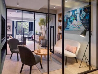 Lounge area at Stey Beijing Wangfujing Hotel, with grey and cream armchairs, a round wooden side table, black-framed glass partitions and a blue artwork on the wall.
