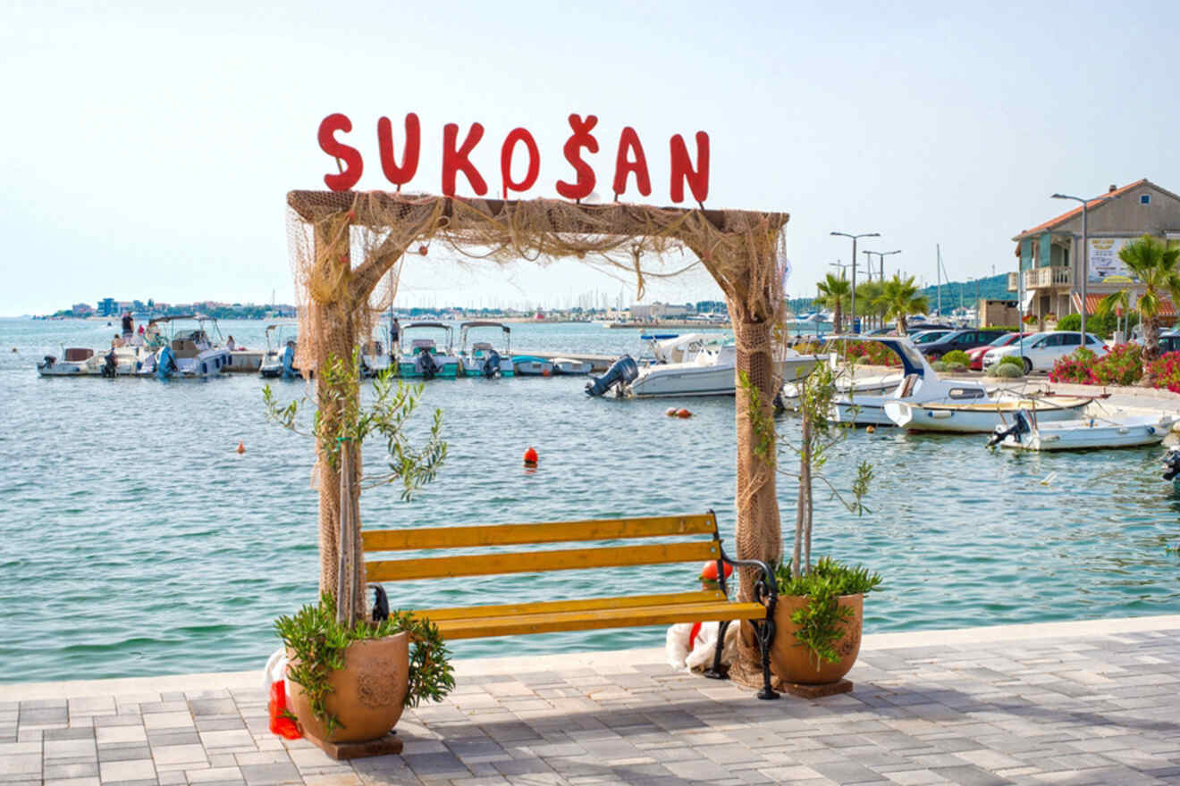 A yellow bench with potted plants on each side sits under an archway with "Sukošan" written on top. Boats are docked in the water behind, and buildings are visible in the background.