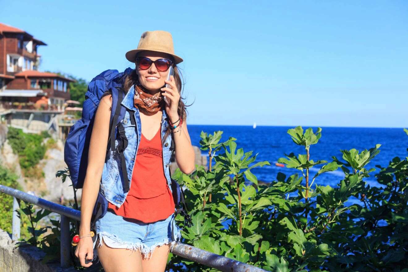 A person with a backpack, hat, and sunglasses stands near greenery by the ocean, talking on a phone. Houses and the sea are visible in the background.