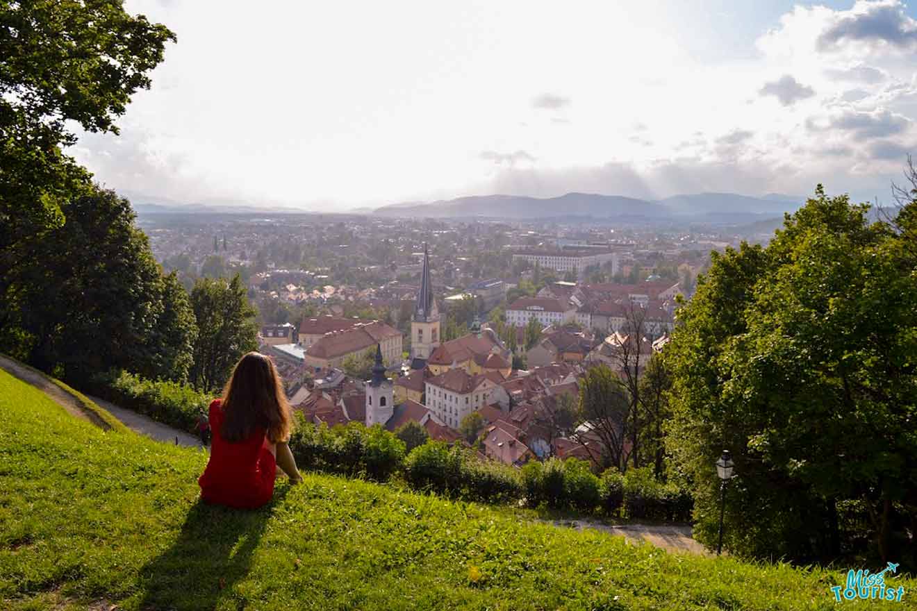 author of the post in red sits on a grassy hill overlooking a picturesque town with red-roofed buildings and a prominent church spire, surrounded by greenery and distant mountains under a partly cloudy sky.