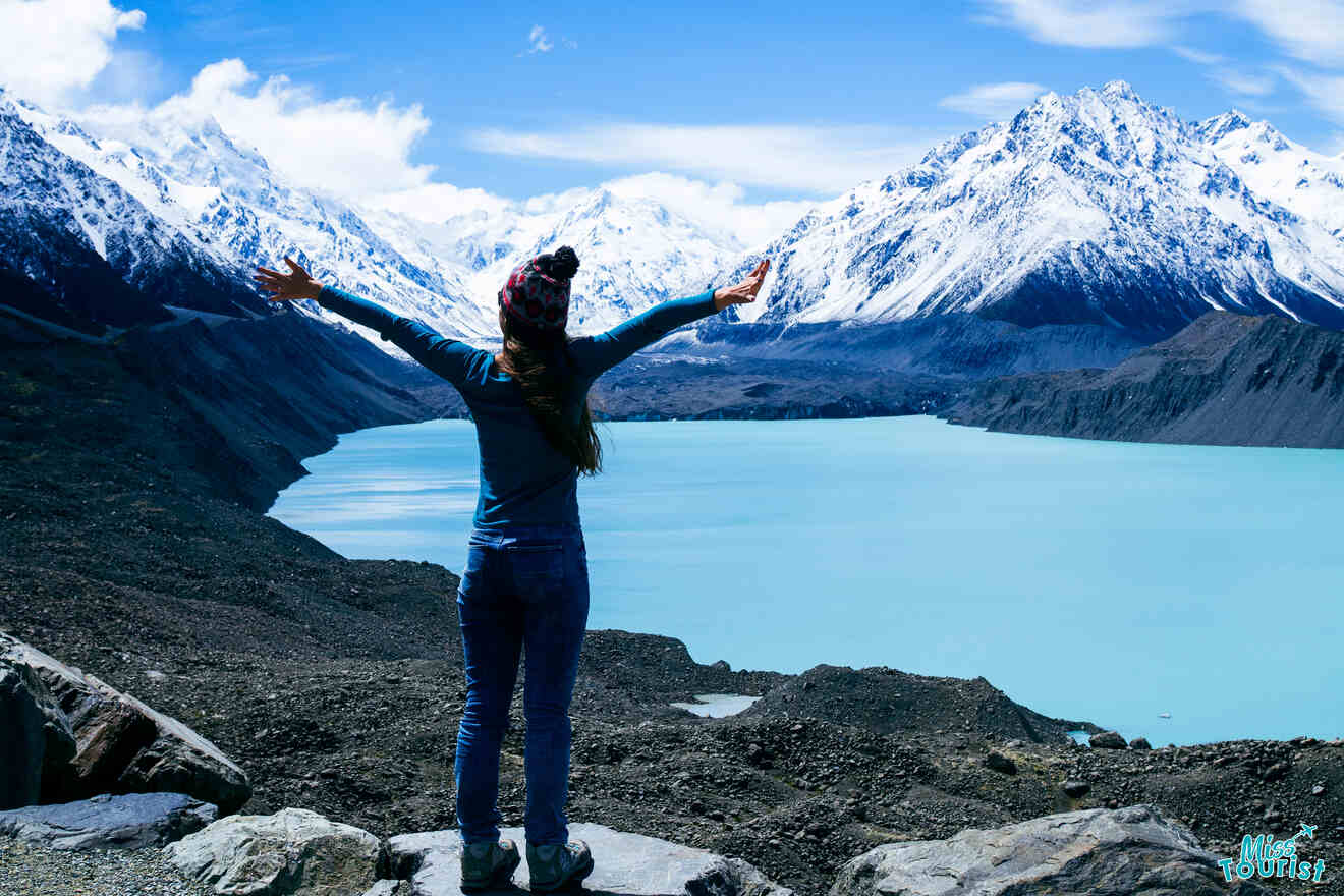 author of the post stands on rocks with arms outstretched, facing a turquoise lake and snow-capped mountains under a blue sky.