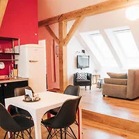 A modern apartment with a dining area featuring a white table and black chairs, a kitchen with red walls, a white fridge, and a living area with a gray couch and TV under large skylight windows.