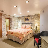 A bedroom with a stone accent wall, a bed with floral bedding, two bedside lamps, a wicker chair, and a small desk under warm ceiling lights.