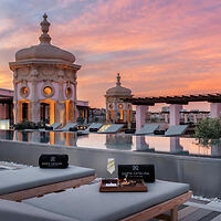 A rooftop pool area with cushioned lounge chairs, ornate architectural structures, and a sunset sky in the background.