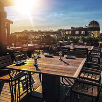 Rooftop terrace at sunset with empty wooden tables and chairs, set with wine glasses and cutlery, overlooking an urban skyline.