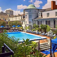 Outdoor swimming pool surrounded by lounge chairs and umbrellas with a historic building in the background under a blue sky.