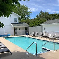 Outdoor swimming pool with lounge chairs lined up along the sides, white buildings in the background, and trees providing partial shade. The sky is clear with some clouds.