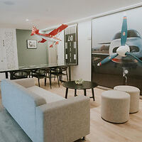 A modern living room with an aviation theme, featuring a gray couch, two round ottomans, a black coffee table, wall art of airplane propellers, and a suspended model aircraft.