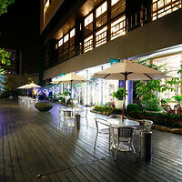Outdoor dining area at night with metal tables and chairs, white umbrellas, string lights, and lush greenery next to a modern building.