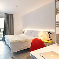 A modern hotel room with a neatly made double bed, a red chair at a desk with a closed laptop on it, soft lighting, and a large window with gray curtains.