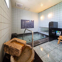 A small indoor bathhouse featuring a tiled soaking tub, wooden stools, and washing stations. Stone basins are in the foreground. The walls are tiled with a large sign above the tub.