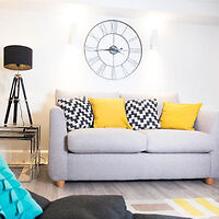 A brightly lit living room with a gray sofa, three yellow and patterned throw pillows, a black floor lamp, a small side table, and a large wall clock above the sofa.