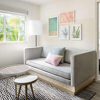 A modern living room with a gray couch, a round coffee table, a tall floor lamp, and wall art above the couch. A window with a view of greenery provides natural light. A patterned rug covers the floor.