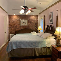 A bedroom with a large bed, two bedside tables with lamps, a brick accent wall, framed artwork, and a ceiling fan. The room has hardwood floors and soft lighting.