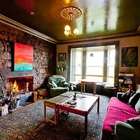 A cozy living room with stone walls, a lit fireplace, and a mix of furniture including armchairs and a sofa. The ceiling is reflective and a large window allows natural light to enter the space.