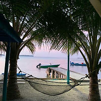 A hammock hangs between two palm trees, facing a wooden pier and a green boat on a tranquil body of water at sunset. Other boats are visible in the background.