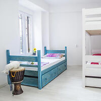 A bright bedroom with a single blue bed, a white bunk bed, a drum beside the blue bed, and a window allowing natural light. Two mattresses have pillows and minimal bedding.
