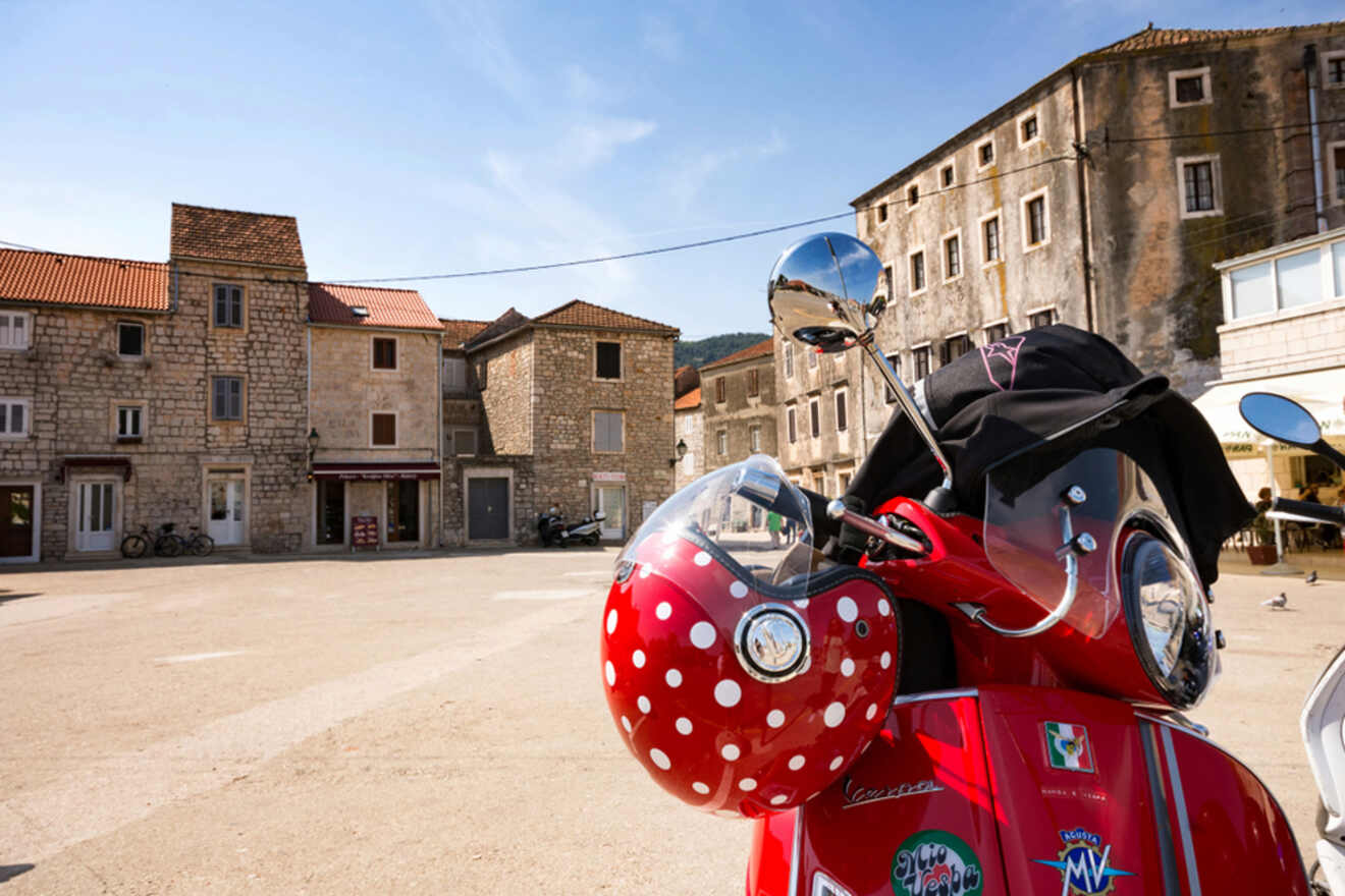 A red Vespa scooter with a polka dot helmet is parked in a sunny, empty town square surrounded by stone buildings.