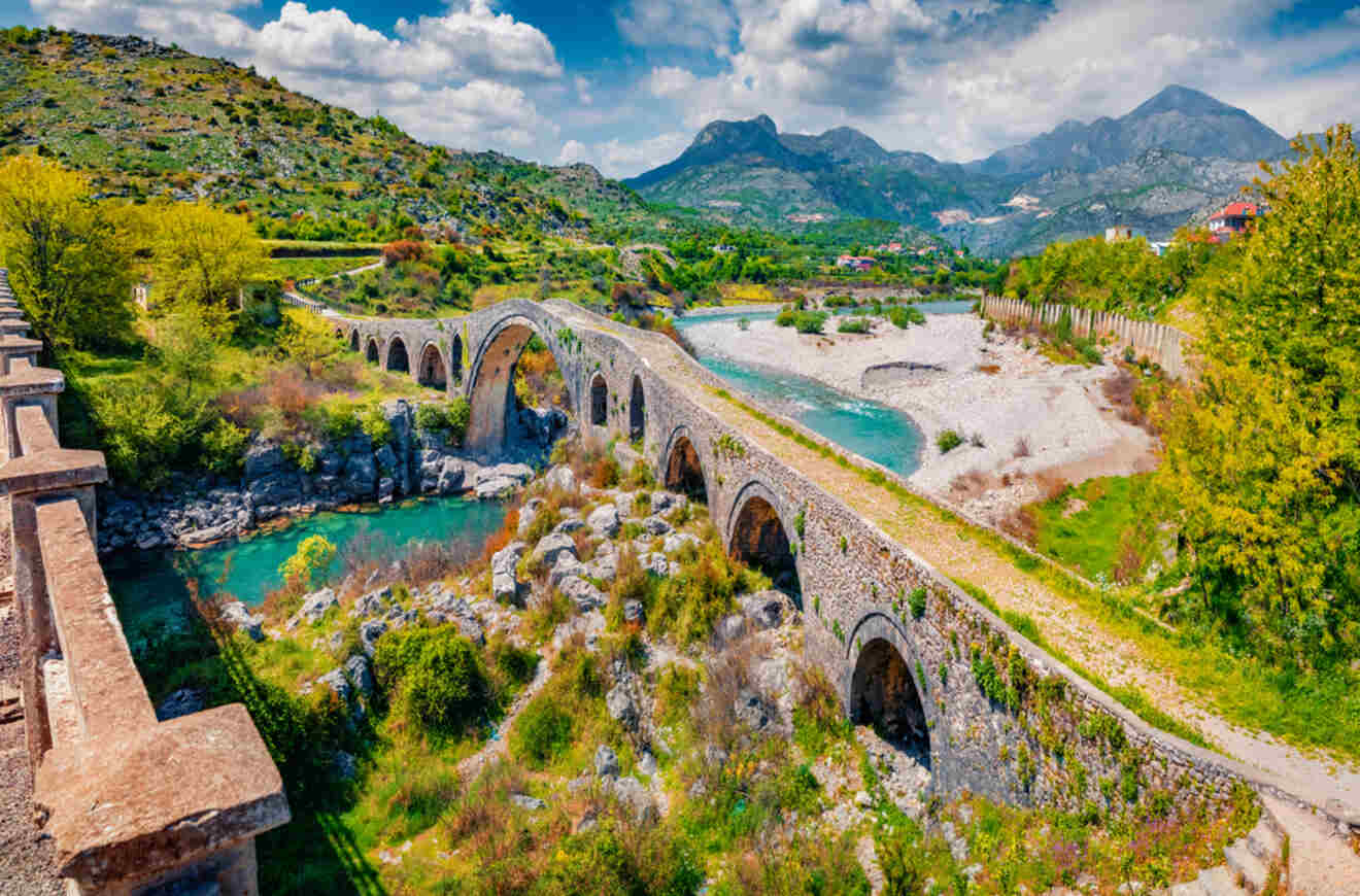 The historic Mes Bridge spanning over a turquoise river, set against a backdrop of lush green hills and mountains.
