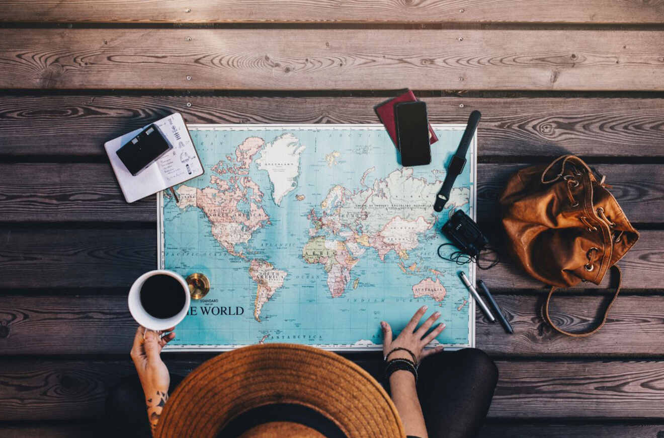 A person sits with a cup of coffee, a hat, and travel accessories on a wooden surface with a world map laid out in front.
