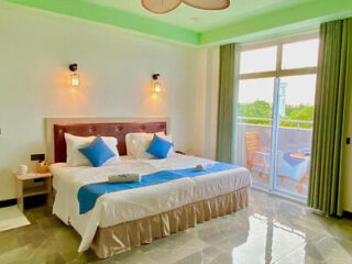 A hotel room with a large bed, white and blue bedding, two bedside tables, wall sconces, and a glass door leading to a balcony with two wooden chairs and a scenic view outside.