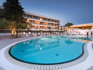 A serene outdoor pool with lounge chairs and a large tree nearby. The pool area is bordered by a multi-story building with illuminated balconies at dusk.