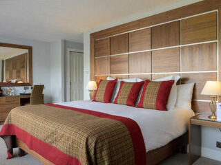 A hotel room features a double bed with plaid bedding and red accents, two bedside tables with lamps, a large mirror, and a desk with a chair. The headboard has a geometric wooden design.