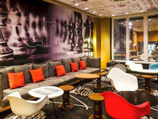 A modern lounge area with a gray sectional sofa adorned with red pillows, various chairs, and tables, and a wall mural featuring chess pieces. The area is well-lit with contemporary décor.