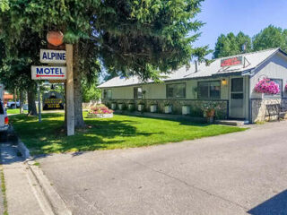 A small motel with a sign reading "Alpine Motel," surrounded by greenery and flowers. There is a parking lot partially visible on the left side of the image.