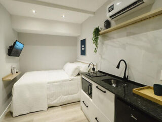 A compact modern studio apartment with a bed, wall-mounted TV, small kitchen area with sink, cabinets, and a countertop. The room is decorated with a framed artwork and a hanging plant.